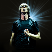 Image of Richard Ashcroft in a black t-shirt wearing sunglasses and holding a microphone, set against a static black background backlit by a bright white spotlight